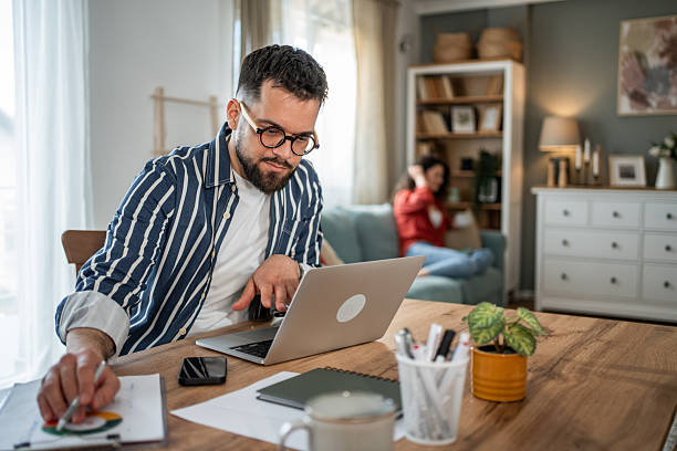 istock Focused businessman working from home analyzing charts and using laptop while woman relaxing on sofa 2214872288