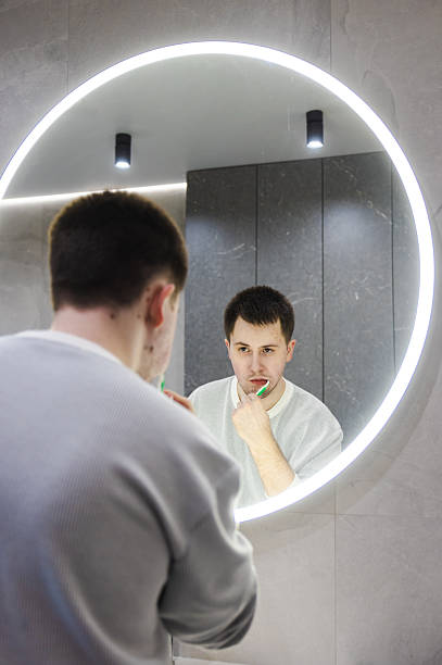 istock Young man in a light sweater brushes his teeth while looking in a round LED-lit mirror, maintaining his morning routine in a modern bathroom. 2214878799