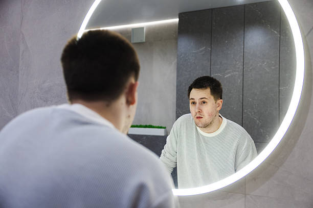 istock Sleepy and surprised young man looks at his reflection in a round illuminated mirror, standing in a modern bathroom during a morning routine. 2214879013
