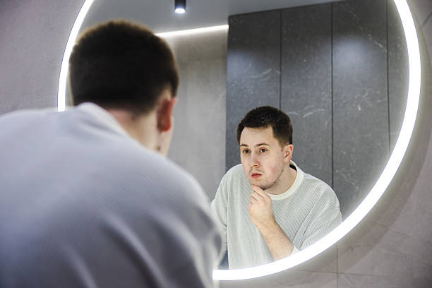 istock A concerned young man examines his face closely in a round illuminated mirror, touching his chin during his morning grooming routine in a modern bathroom. 2214879302