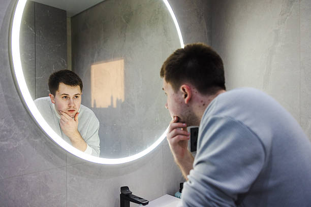 istock Man in a light sweater leans over a bathroom sink, thoughtfully examining his reflection in a round LED-lit mirror during his morning routine. 2214882250