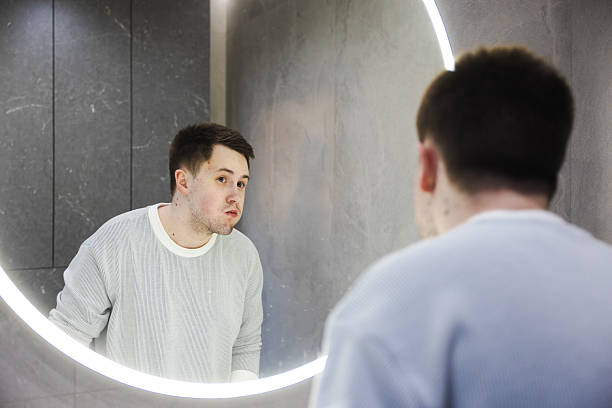 istock Man in a light sweater rinses his mouth while looking into a round illuminated bathroom mirror, finishing his morning dental routine. 2214882388