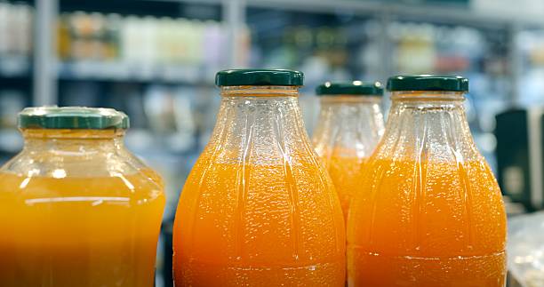 Multiple glass bottles filled with orange juice are neatly arranged on a supermarket shelf, creating a vibrant display against a backdrop of other products