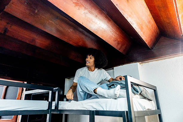 Young man with curly hair sitting comfortably on a top bunk bed in a cozy room with wooden ceiling beams. He is wearing a light-colored t-shirt, jeans, and socks, looking to the side with a relaxed expression.