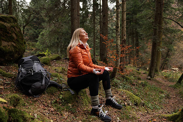 A woman in an orange jacket meditates calmly outdoors, enjoying nature's tranquility. Autumn leaves surround her in a serene forest environment.
