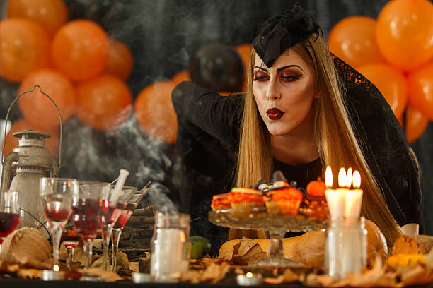 Woman dressed in a gothic costume blowing a lit candle surrounded by Halloween-themed decorations and an eerie ambiance characterized by orange balloons, spooky props, and festive treats arranged on a table.