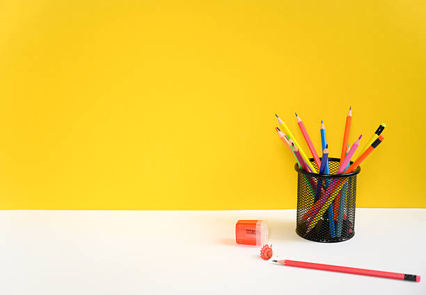Multicolored pencils in black holder with sharpener placed on minimal desk with yellow wall. Ideal for school design, education themes, creative compositions, school promotions and  kids learning.
