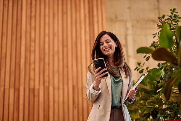 Businesswoman using smartphone and holding clipboard is smiling in modern office