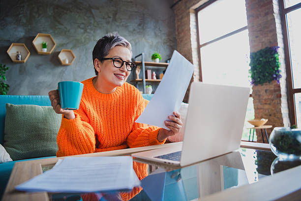 istock Photo of smiling positive elderly lady wear orange pullover working modern device reading report indoors apartment room 2214949773