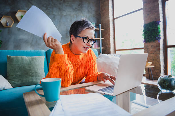 istock Photo of positive happy elderly lady wear orange pullover reading modern device writing report indoors apartment room 2214949900