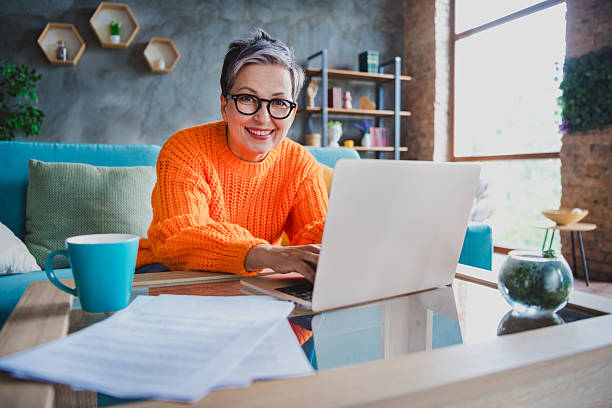 istock Photo of happy positive elderly lady wear orange pullover texting emails modern device indoors apartment room 2214950128