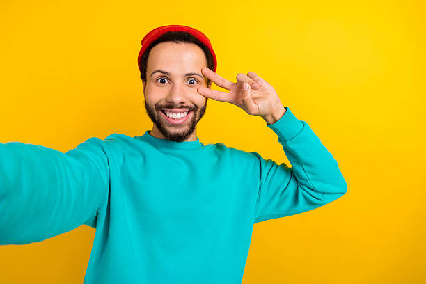 istock Portrait of positive funky man with beard dressed turquoise pullover making selfie show v-sign on eye isolated on yellow color background 2214950890