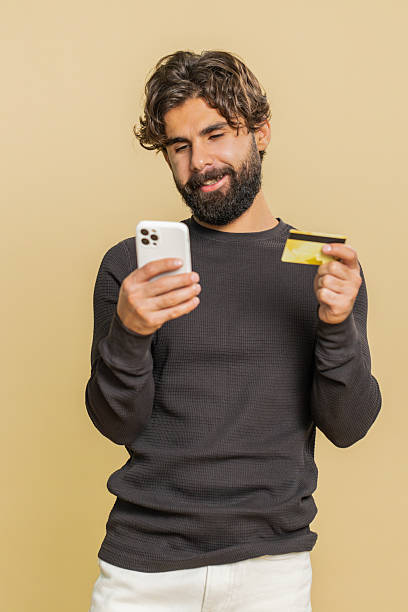 istock Happy young man using credit card and smartphone while transferring money against beige background 2214951156
