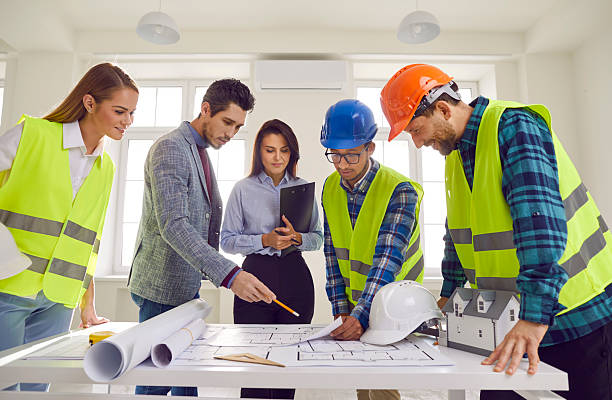 istock Construction team standing around office table and looking at paper blueprint plans 2214968937