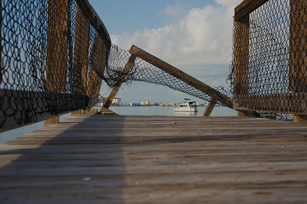 Damaged Wooden Pier Overlooking Waterfront With Urban Skyline and a white Docked Boat. Gulfport, FL bent netting frames a peaceful waterfront view featuring a white boat and urban skyline in the distance under a cloudy morning sky, evoking tranquility and reflection. gulfport florida photos stock pictures, royalty-free photos & images
