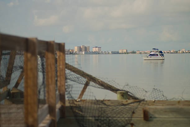 Damaged Wooden Pier Overlooking Waterfront With Urban Skyline and a white Docked Boat. Gulfport, FL bent netting frames a peaceful waterfront view featuring a white boat and urban skyline in the distance under a cloudy morning sky, evoking tranquility and reflection. gulfport florida photos stock pictures, royalty-free photos & images