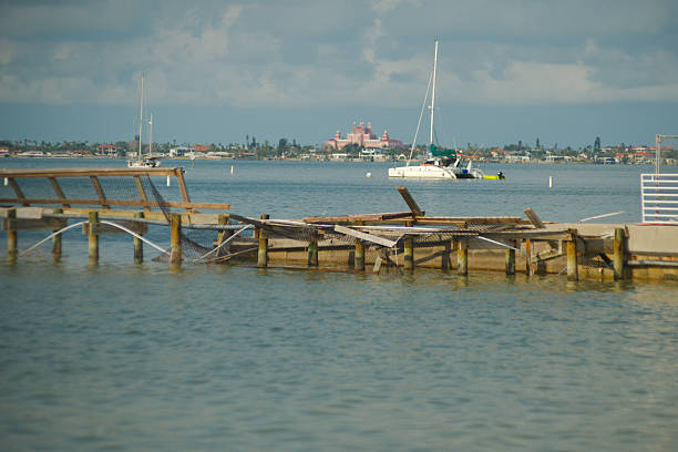 Scenic View of Waterfront with Boats and Distant Historic Pink Landmark Gulfport, FL tranquil coastal scene featuring a damaged wooden pier, anchored sailboats, calm water, and the pink Don Ceasar hotel building in the distant shoreline under a cloudy sky. gulfport florida photos stock pictures, royalty-free photos & images