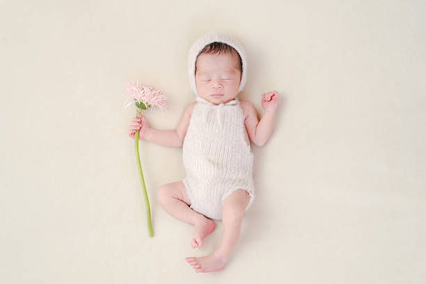 A 7-day-old Japanese newborn baby, in a pink romper and hat, holds a pink gerbera while resting on a white background. This soft image captures the beauty and innocence of early life.