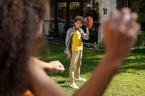 Child standing on green lawn under sunlight, holding baseball glove and smiling at camera surrounded by house and trees looking at camera