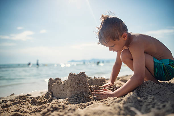 A young child crouches in the warm beach sand, using hands to shape his creation. \nShot with Nikon D800 on a sunny summer vacations day.