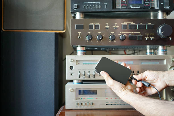 One hand holding a USB charging cable or charger wire and plugging it in or holding close to a portable music radio device with steel aerial antenna over yellow t shirt and horizontal gray color background