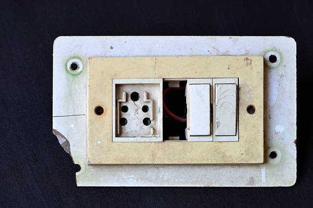 Rusty rustic weathered distressed damaged hazardous run down broken messy dirty cream electrical board plate with electricity wire or cable, three pin electric socket, two switches installed and one missing part making recyclable waste on black background with copy space.