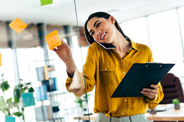 istock Qualified Businesswoman in Casual Office Setting Analyzing Notes on a Glass Wall While Holding a Clipboard 2215000405