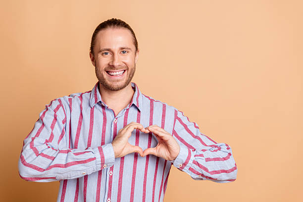 istock Smiling young man with neatly groomed beard making heart shape with hands on beige background wearing striped casual shirt 2215000899