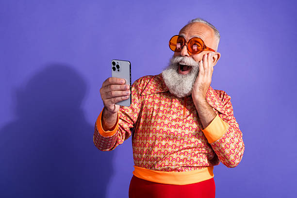 istock Happy senior man in retro themed clothing expressing excitement while using smartphone against vibrant purple background 2215002553