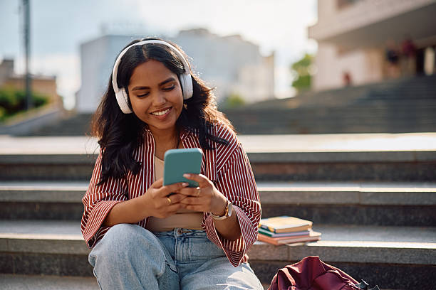 Happy Indian student using her cell phone while listening music over headphones at campus.