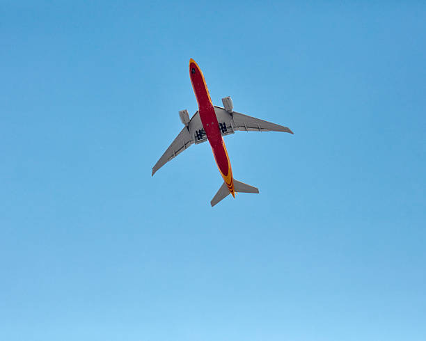a wide-body airliner seen from below, flying in a clear, cloudless blue sky. The airplane is predominantly yellow, with a bright red stripe running along the body of the aircraft. The retracted landing gear is slightly visible. The tail of the airplane is tilted slightly downward, suggesting that it is on approach to land.