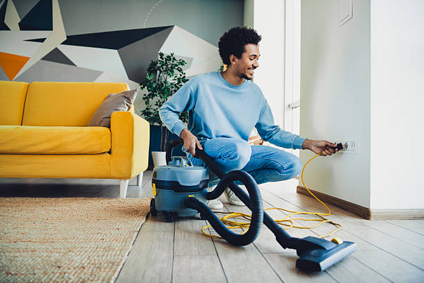 istock Smiling young man plugging in a vacuum cleaner while preparing to clean his cozy and stylish living room on a sunny day 2215004056