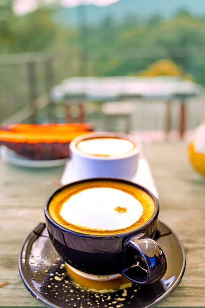 Food Series : Hot coffee in black ceramic cup with nature background. Close up shot of hot latte coffee. Cappuccino with hot milk cream. Two cups of hot coffee on the table.