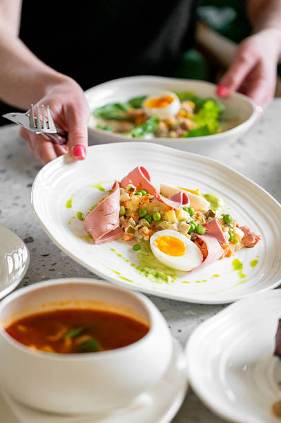 istock A plate of food featuring sliced ham, a boiled egg, peas, and a vegetable mix, garnished with green sauce, served alongside a bowl of soup 2215010247