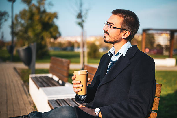 Stylish male entrepreneur enjoying sipping coffee and sitting on bench in city park, representing work-life balance and relaxation.