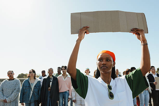 A group of protestors gathered outdoors, with a woman holding up a blank cardboard sign, symbolizing advocacy and expression for a cause, set against a sunny sky backdrop.