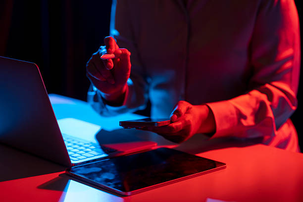 istock Businesswoman holding a cellphone while touching or pointing at something with a marker. Laptop computer and tablet on the table. Mockup and copy space. Business and technology concept. Illuminated with blue and red light 2215039199