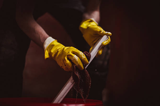 One young unrecognizable Caucasian man in yellow gloves washes part of plastic window frame with rag and flowing water, stands bent over in house, bottom side view close-up with selective focus. House renovation and window frame washing concept.