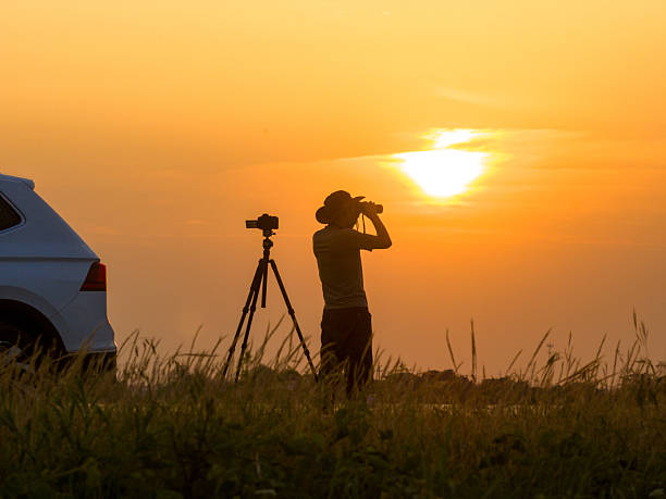 An Asian man using a telescope to watch birds outdoors, sunset silhouette