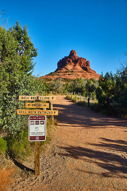 Path leading to bell-shaped rock formation on blue sky day with green brush and leafless tree.