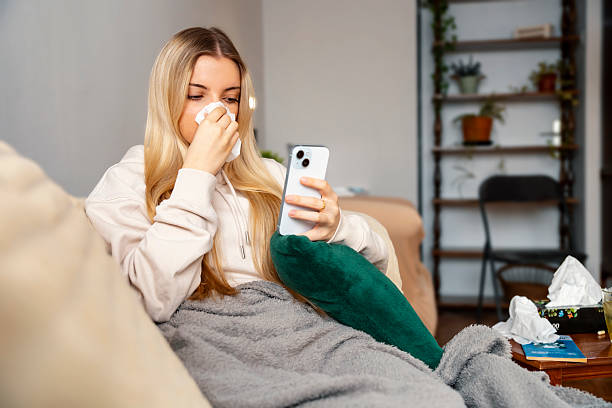istock Young woman using smartphone while blowing her nose with a tissue 2215056973