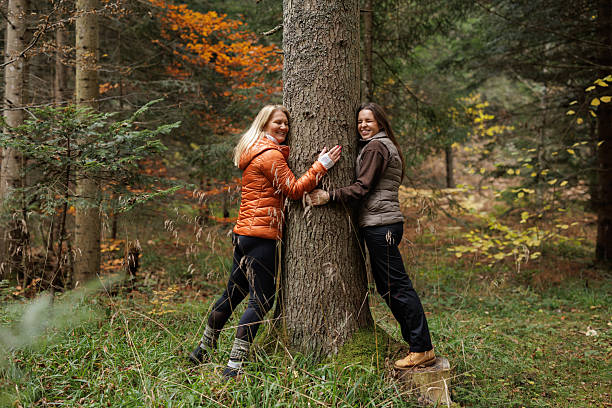 Two women are seen smiling and hugging a tree in a vibrant autumn forest, emphasizing appreciation and harmony with nature.