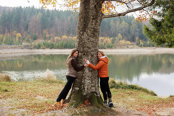 Two women embrace a tree by a serene lake surrounded by forest hues of autumn, reflecting nature's tranquility and connection to the environment. The peaceful scene evokes relaxation and outdoor enjoyment.