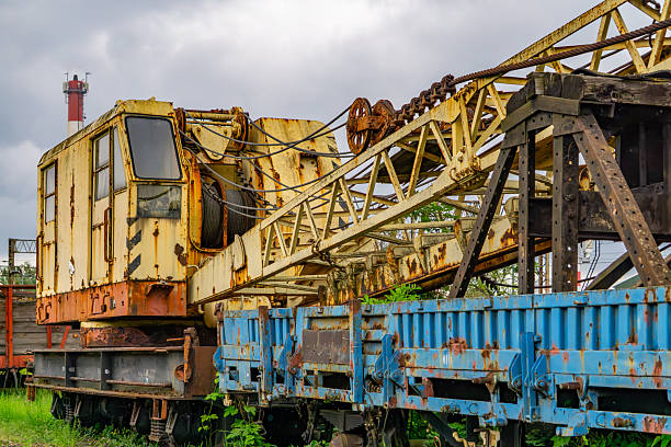 Rusty vintage railway crane mounted on a flatbed railcar, featuring a long boom arm and an operator's cab. The machine shows signs of heavy use and corrosion, set in an industrial railway environment with an abandoned feel.