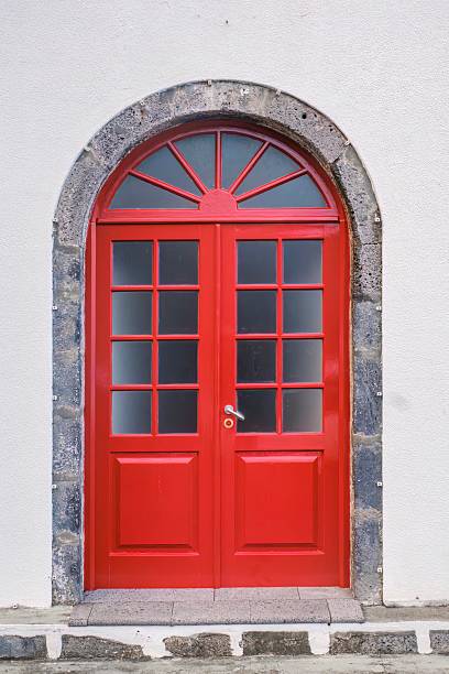 Vibrant Red Double Door with Arched Stone Frame. Eye-catching bright red double doors with glass panes set within a rustic arched frame made of grey stone against a light wall