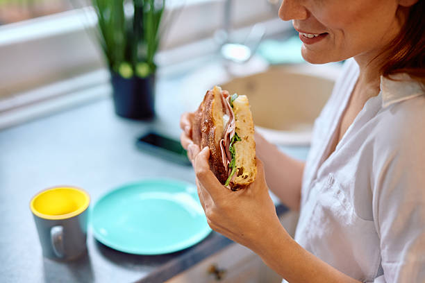 Close-up of a young adult woman holding a mouthwatering sandwich, preparing to take a satisfying bite, while surrounded by a stylish, modern kitchen filled with natural light