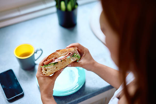 Woman enjoying a delicious breakfast at home, holding a fresh sandwich filled with mortadella, cheese, and arugula, while sipping coffee at the table by the window