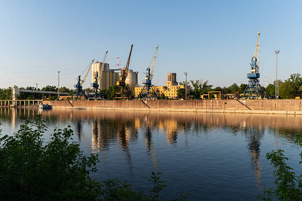 A series of large industrial cranes line a riverside dock, backed by towering silos and warehouse structures. The calm river reflects the golden light of the setting sun, adding a warm hue to the industrial scene.