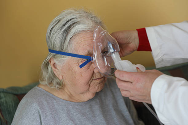 An older woman with gray hair, wearing an oxygen mask attached by blue bands around her head, receives breathing therapy using an inhaler or nebulizer. She is wearing a blue shirt and sitting in a comfortable living room, with plants and a table lamp in the background. Beside her, a medical worker in a white coat and gloves is helping her hold the breathing apparatus. The look on her face reflects the seriousness of the situation, indicating medical care at home.