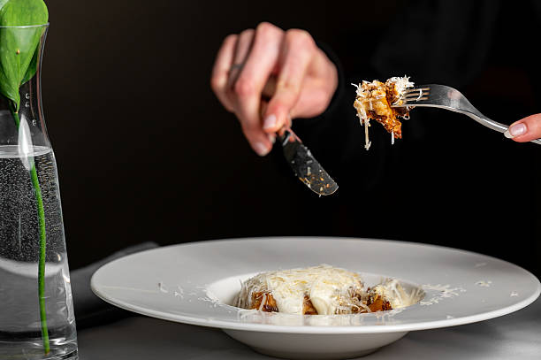 istock A person using fork and knife to eat a dish with melted cheese, served on a white plate. A glass vase with green plant in water is placed beside it. Warm lighting enhances the cozy dining atmosphere 2215091869
